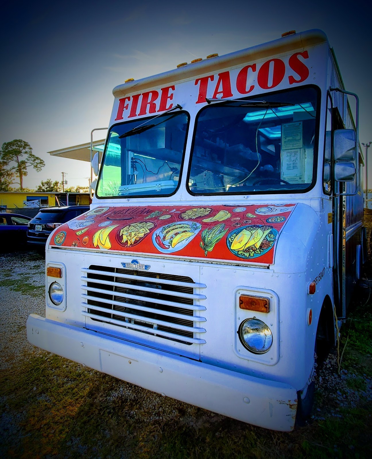 The Fire Tacos food truck at dusk, white panel truck with red FIRE TACOS lettering and a hand-painted hood of illustrated Mexican dishes, parked in Sarasota